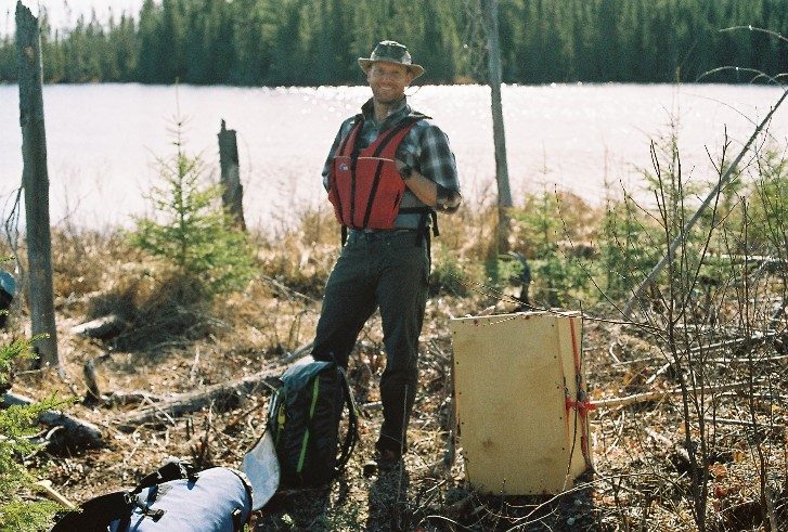 Film photo of a tripper beside his wanigan at the water's edge